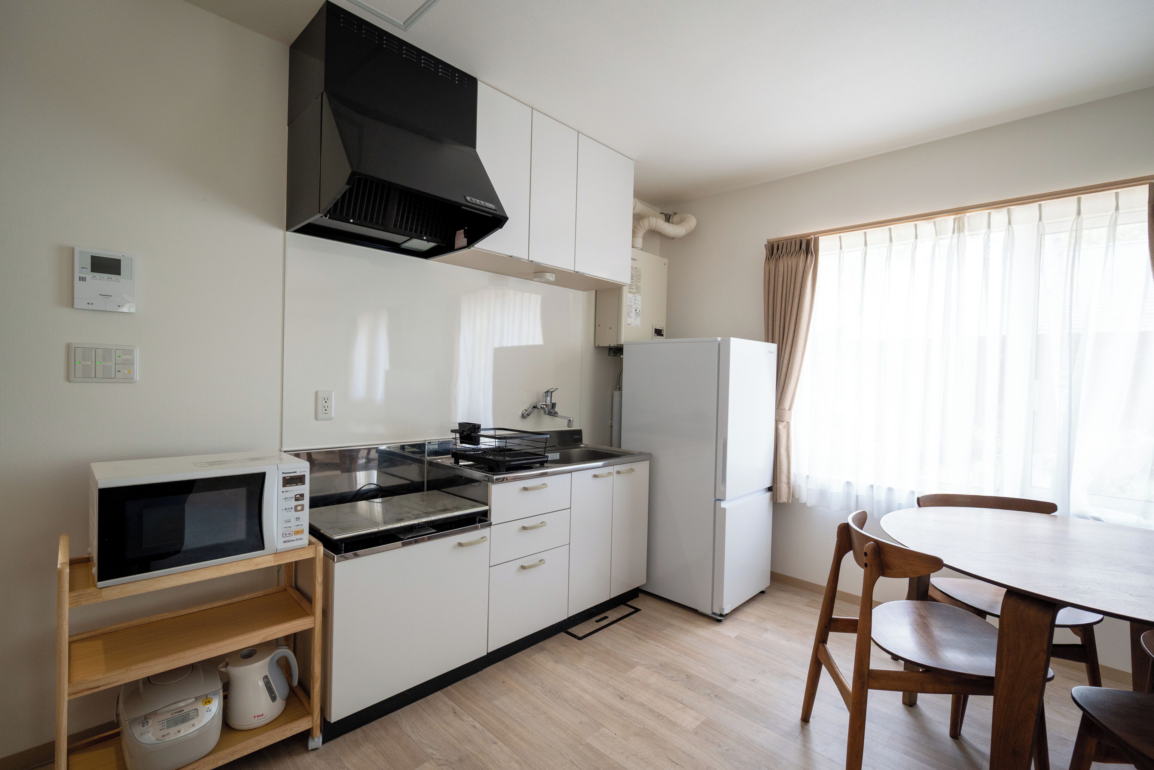 A compact kitchen with a microwave, stove, sink, and refrigerator. There is a wooden table and chairs, and a window with curtains.