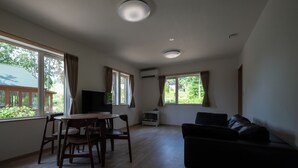 Een kamer met een houten eettafel, stoelen, een zwarte leren bank en grote ramen met gordijnen.