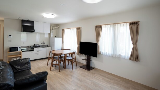 Un moderno salón con televisor de pantalla plana, comedor con mesa y sillas, cocina con alacenas blancas y microondas, y sofá de cuero negro.
