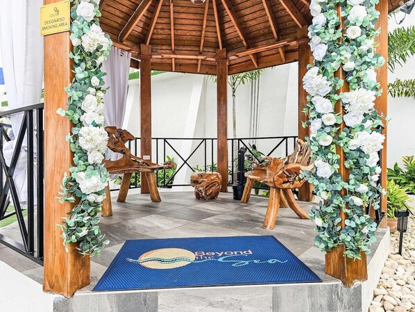 Un kiosque agrémenté de chaises en bois et d’un tapis bleu orné de motifs, entouré de fleurs blanches et de verdure.