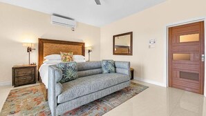 Habitación de hotel con una cama, un sofá gris, un ventilador de techo, una puerta de madera y un aire acondicionado instalado en la pared.