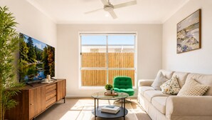 A living room with a TV, a green chair, a sofa, a coffee table, and a window with a view of a fence. - Haus Verde | 4BR Greenbank Hideout in Logan (Greenbank)