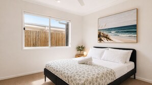 A bedroom with a bed, a window with a view of a beach, a ceiling fan, and a framed picture on the wall. - Haus Verde | 4BR Greenbank Hideout in Logan (Greenbank)