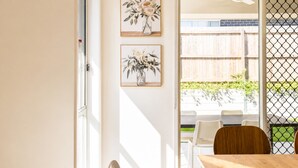A room with a wooden table, two framed floral artworks, a white chair, and a view through a glass door to an outdoor area with a wooden fence and greenery. - Haus Verde | 4BR Greenbank Hideout in Logan (Greenbank)
