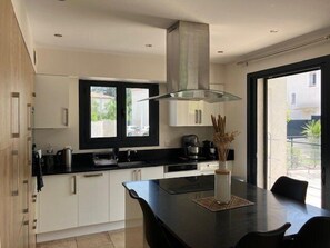 A modern kitchen with white cabinets, a black countertop, a central island, and a large window. - Air-conditioned villa for 8 people with a swimming pool – La Crau (LA CRAU)