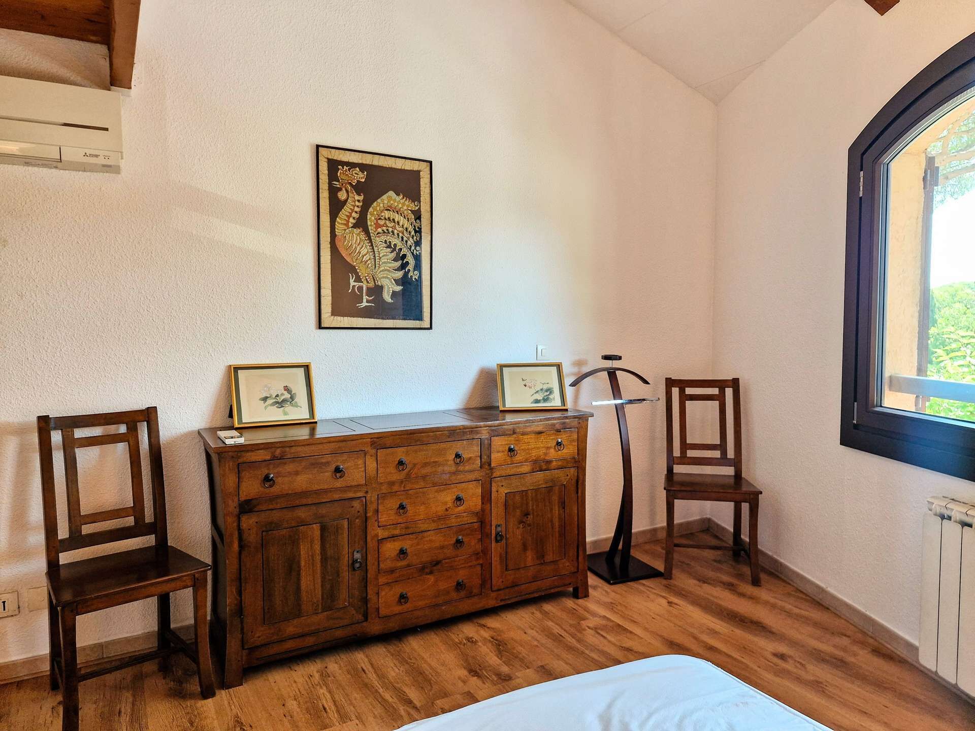 Une pièce avec une commode en bois, deux chaises, un radiateur et une fenêtre donnant sur un paysage agréable.