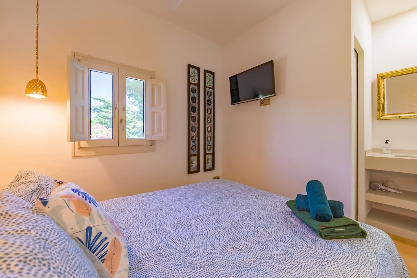 A bedroom with a bed, a TV, a window with shutters, and a shelf with toiletries.