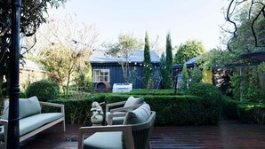 Eine Holzterrasse mit Gartenmöbeln, ein Heckenbusch und im Hintergrund ein Gebäude mit blauem Dach.