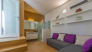 A modern apartment with a staircase, a glass-enclosed shower, a grey sofa with purple cushions, and white shelves with decorative items.
