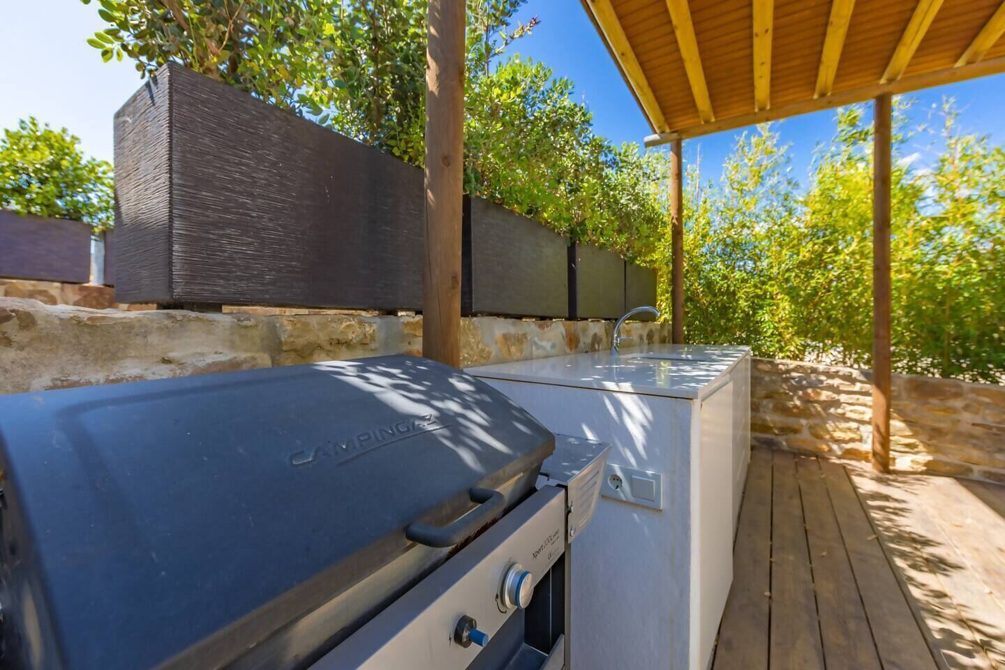 Un espace cuisine extérieur doté d’un barbecue et d’un évier intégrés, entouré d’une terrasse en bois et de murs en pierre.