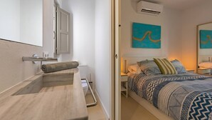A modern bedroom with a built-in bench, a bed with patterned bedding, a bedside table, and a wall-mounted air conditioner.