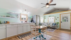 A modern kitchen with white cabinets, a dining table with chairs, a ceiling fan, and a living area with a sofa and a coffee table.