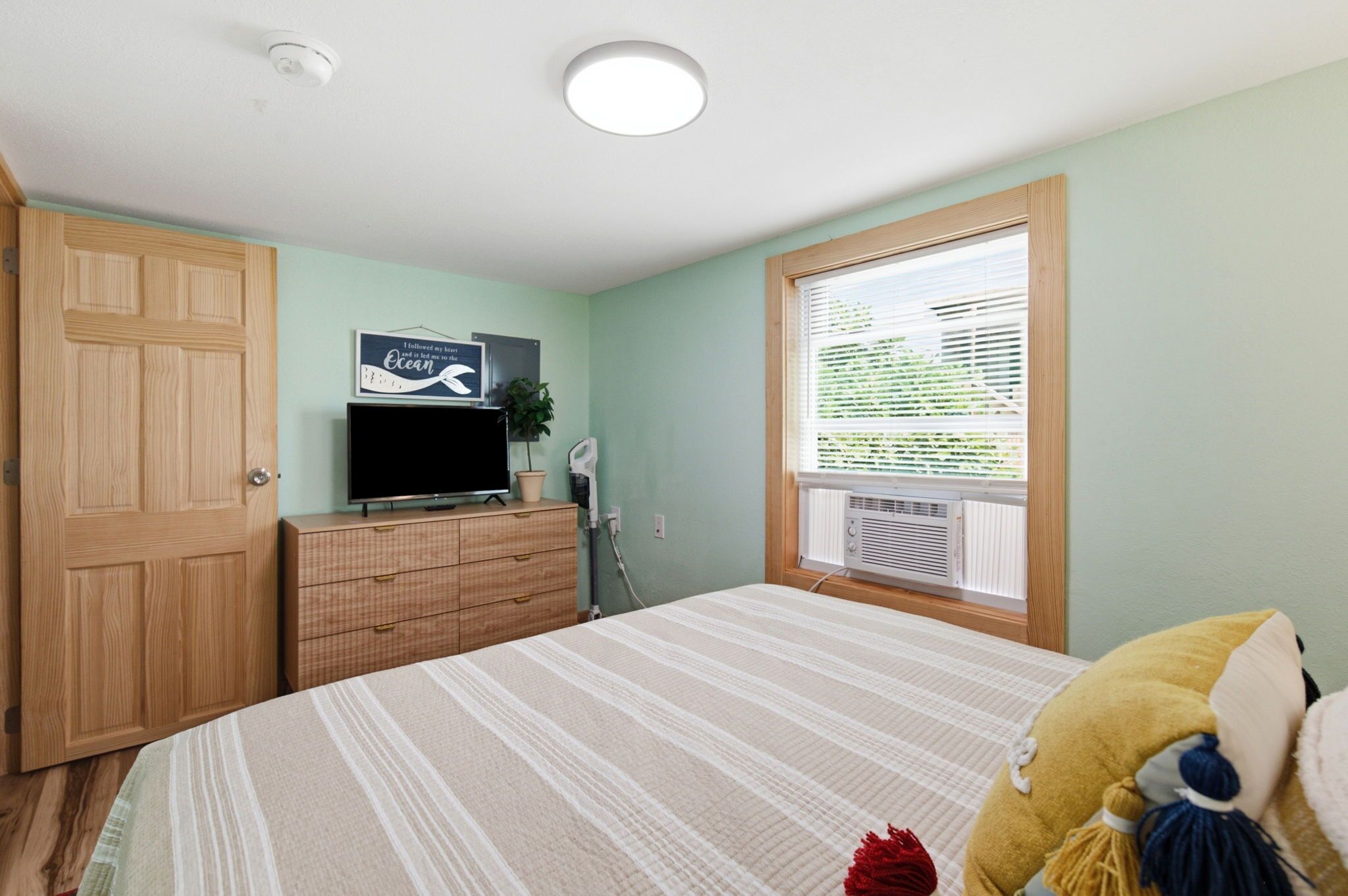 Une chambre à coucher avec un lit, un téléviseur posé sur une commode en bois, une fenêtre avec des stores et une porte.