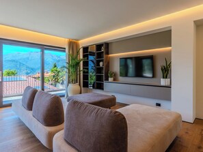 A modern living room with a large window, a flat-screen TV, and a built-in bookshelf. - Vacation home Aura by Interhome (Colico)