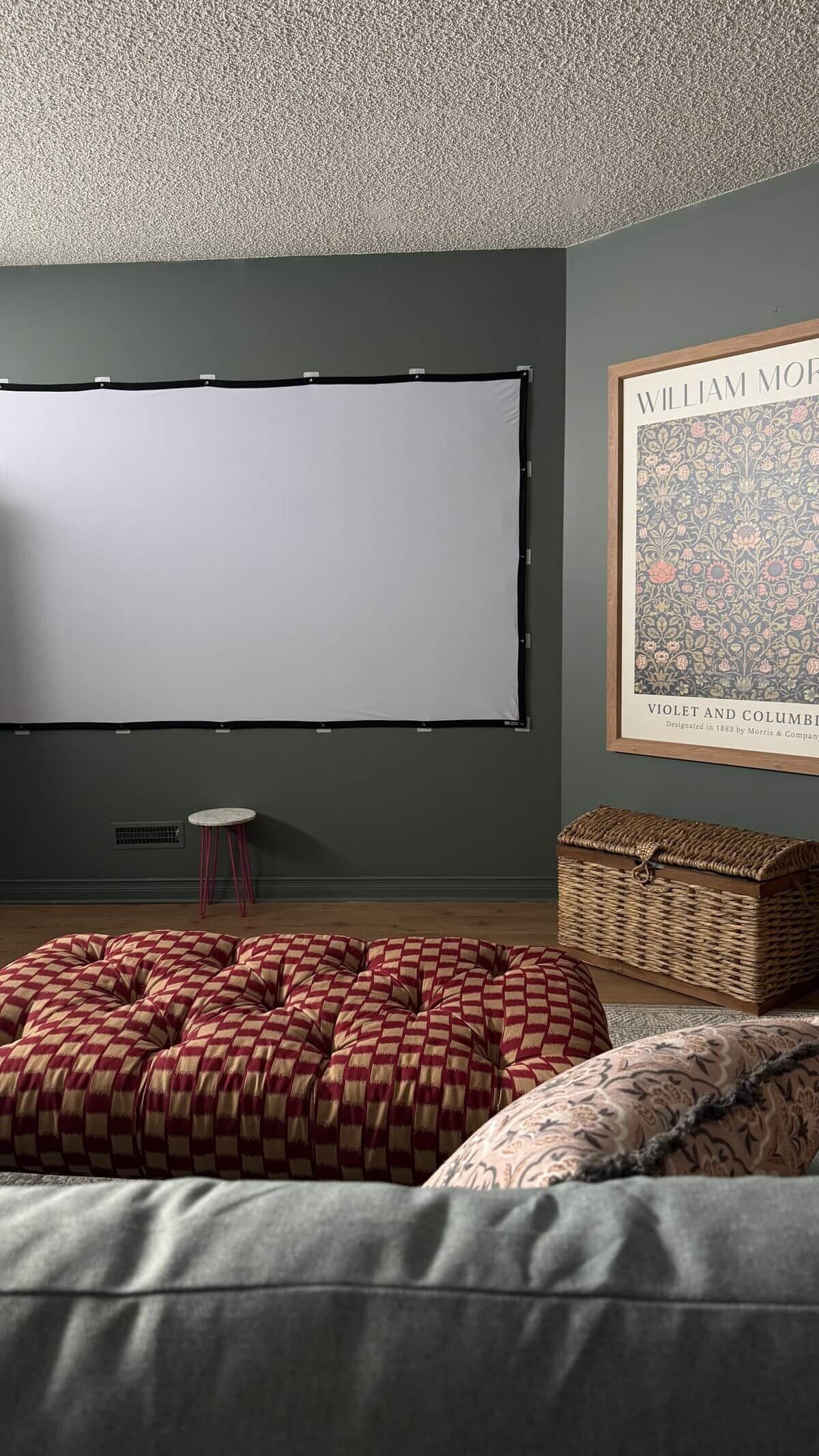Ein Zimmer mit einer großen, leeren Projektionsleinwand, einem gemusterten Hocker, einem Korbwäscher und einem gerahmten Poster an der Wand.