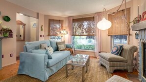 En stue med sofa, lænestol og sofabord, pejs og store vinduer med gardiner.