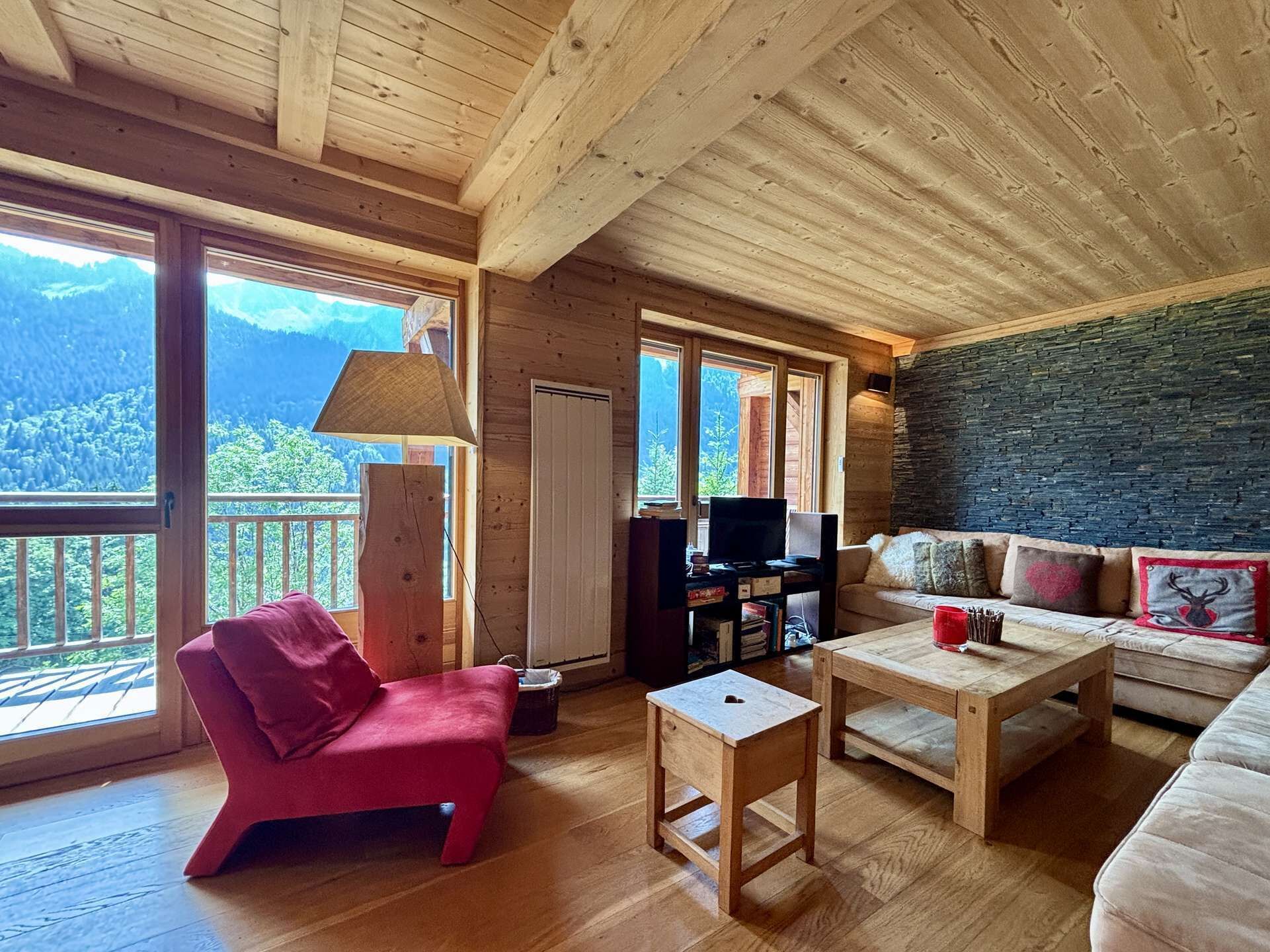 Interior de una cabaña de madera con sofá, una silla roja, una mesa, un televisor y un balcón con vista a la montaña.