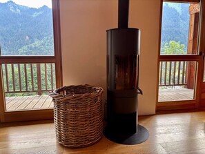 A room with a wood-burning stove, a wicker basket, and a view of a forested mountain through large windows. - Chalet 4 Jillou - Jacuzzi  et  Alpine panorama (LA CHAPELLE D ABONDANCE)