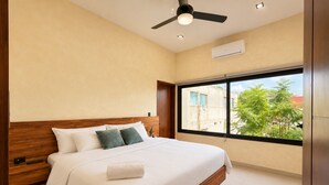 A modern hotel room with a large bed, wooden headboard, a ceiling fan, and a window with a city view.