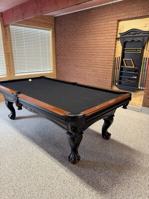 A pool table with a wooden frame and black felt, a brick wall, and a window with blinds.
