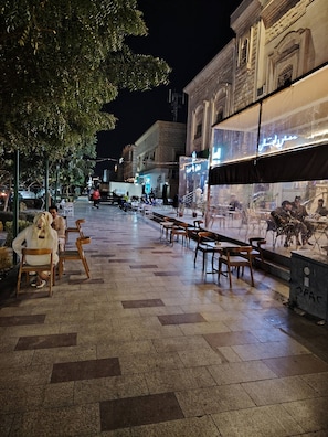 An outdoor seating area with tables and chairs, a tree, and a building with a classical architectural style in the background. - Khair Jewaar Big Ground Apart (Madinah)