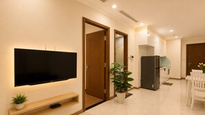 Un salón moderno con una televisión de pantalla plana, puertas de madera, una planta en maceta, un comedor con mesa y sillas blancas, y una cocina con refrigerador.
