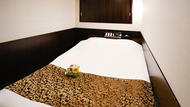 A hotel room with a bed, a dark wooden wardrobe, a small table with a telephone and a clock, and a decorative item on the bed. - ikebukuroCentralhotel (Tokyo)