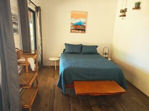 Un dormitorio con cama, una mesita, una silla, un ventilador de pared y un cuadro.
