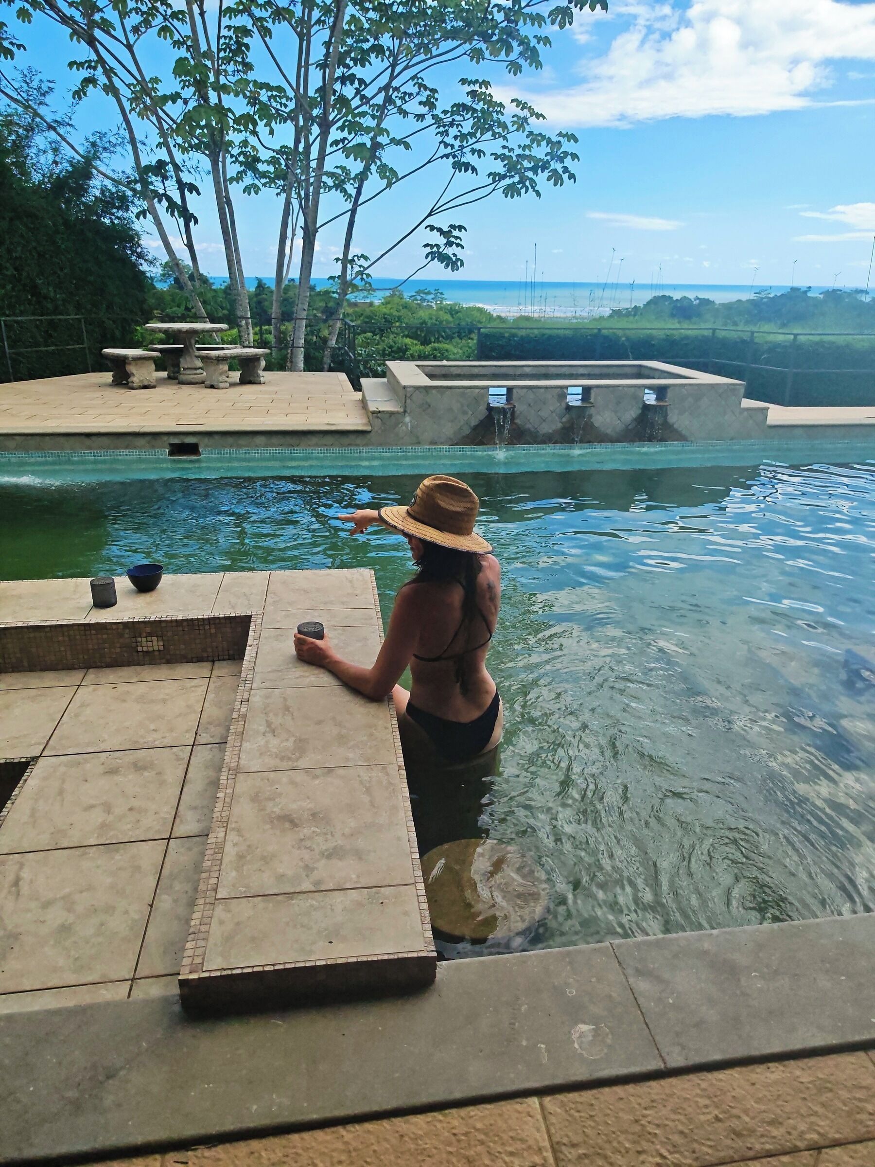A person standing at the edge of a swimming pool with a stone deck and a stone table with chairs.