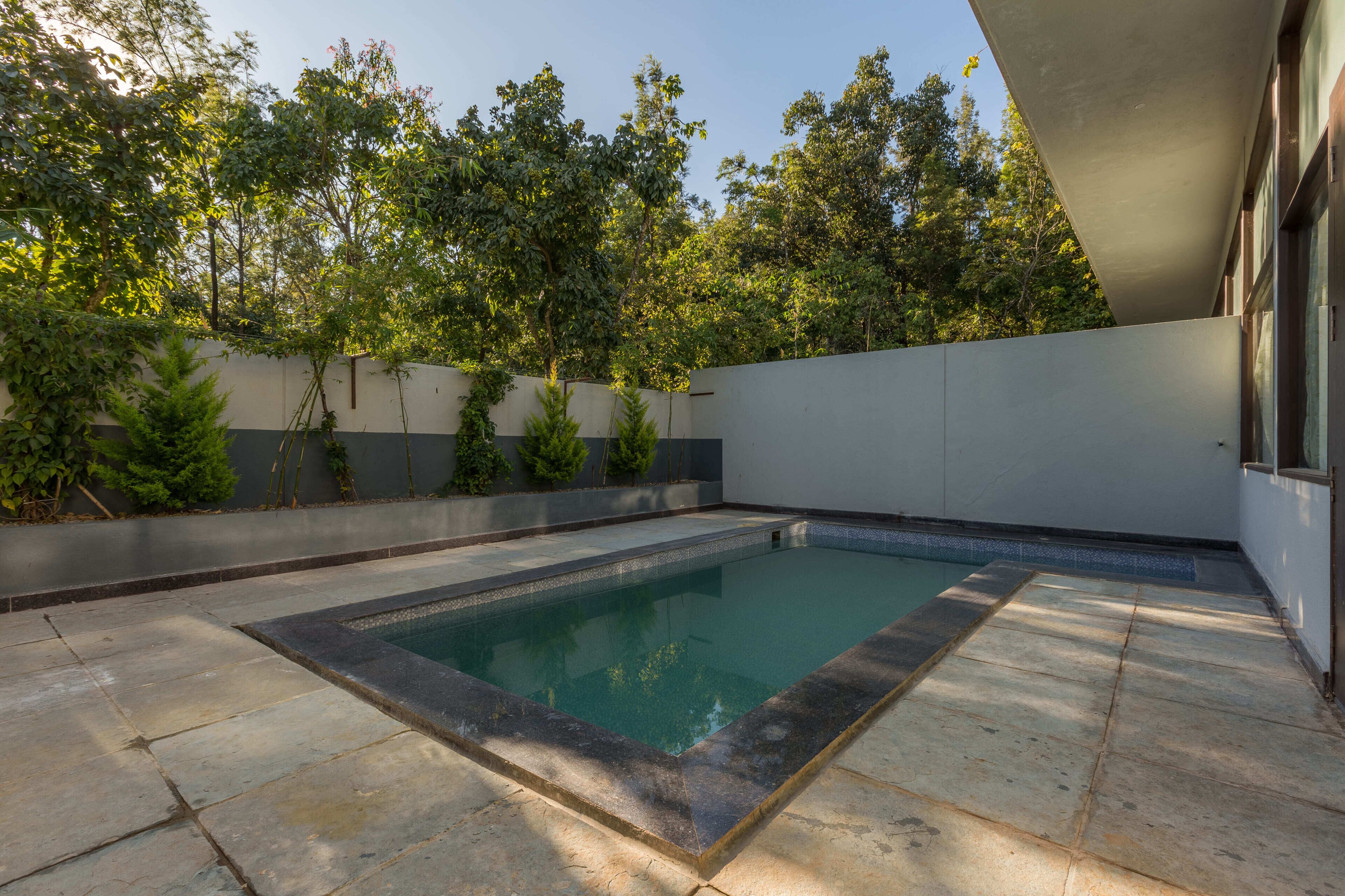 Piscina rectangular con borde de piedra, rodeada por un patio empedrado y un muro bajo con vegetación.