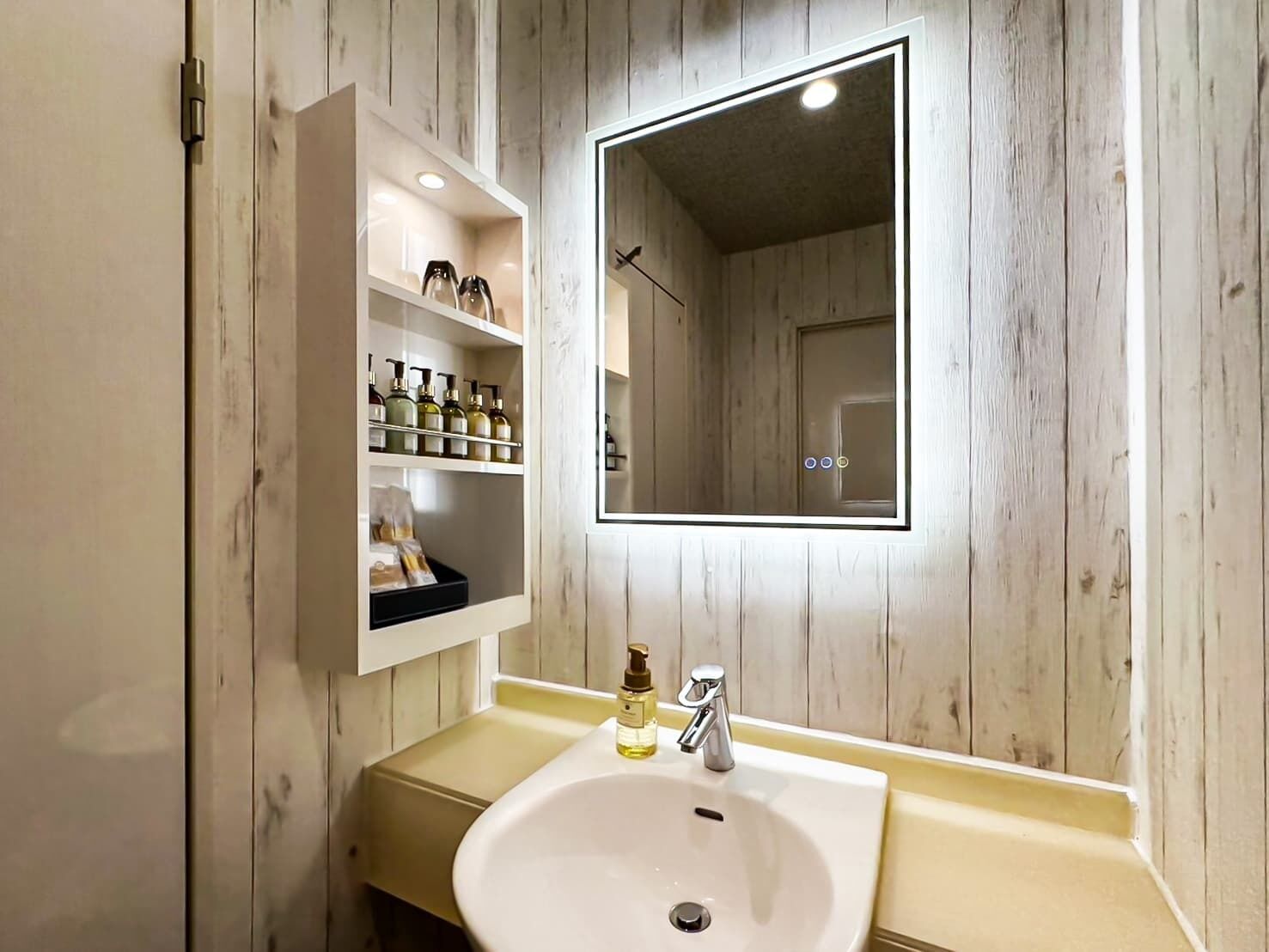 Un baño con lavamanos blanco, un espejo y un estante con artículos de tocador.
