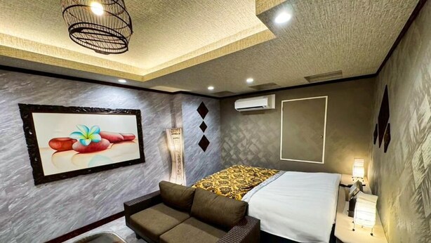 Une chambre d’hôtel comprenant un lit, un canapé, un fauteuil en osier, un climatiseur fixé au mur et un tableau encadré.
