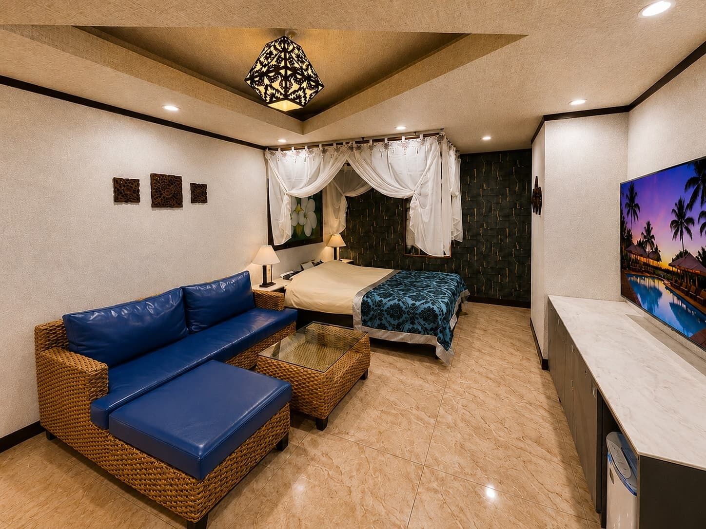 Une chambre d’hôtel avec un lit, un canapé bleu, une petite table, un téléviseur et un tableau représentant un paysage tropical.