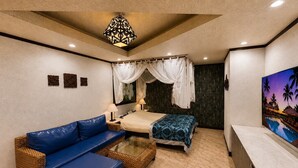 Una habitación de hotel con una cama, un sofá azul, una mesita, un televisor y un cuadro de una escena tropical.