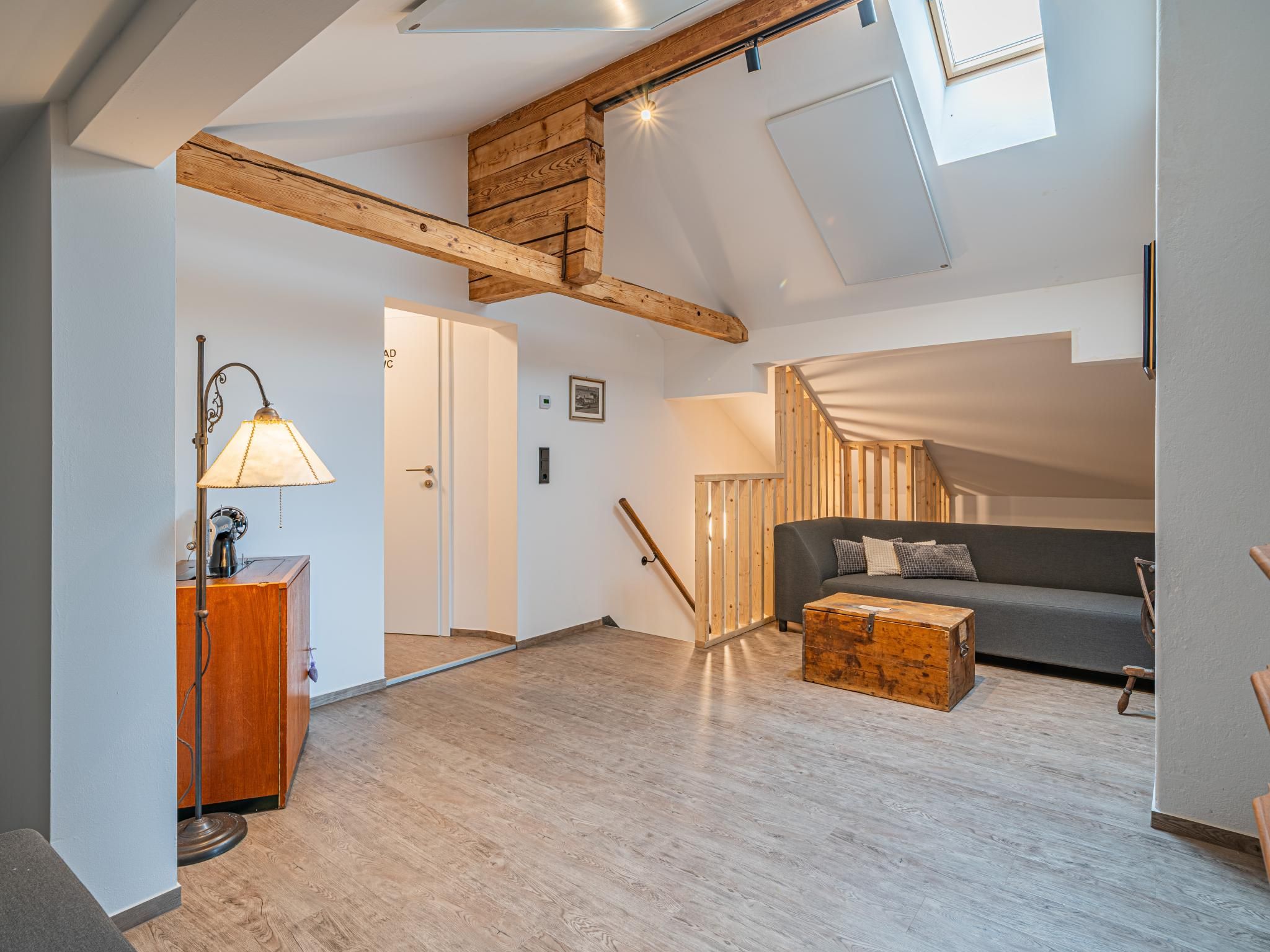 Un loft spacieux avec un plafond haut, des poutres apparentes, un escalier, un canapé, une table basse et un bureau avec une lampe.