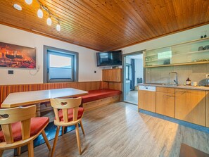 A cozy cabin interior with a wooden ceiling, a dining table with chairs, a bench, a television, and a kitchen area.