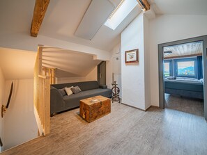 A loft-style room with a loft bed, a grey sofa, a wooden chest, and a spinning wheel.