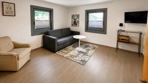 A modern living room with a sofa, a small round table, a television, and two windows with blinds.