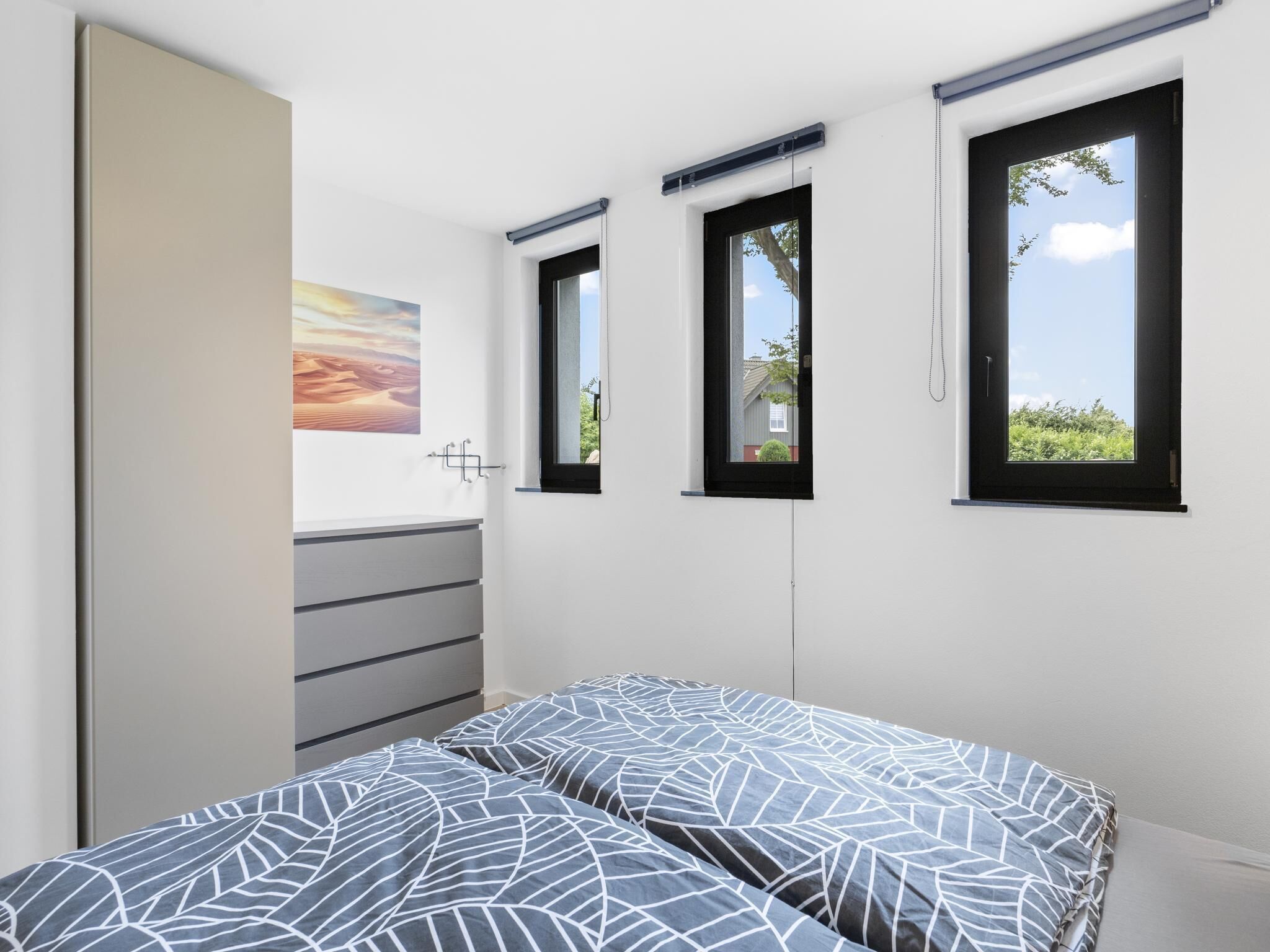 Un dormitorio con una cama, un aparador, un cuadro y tres ventanas.