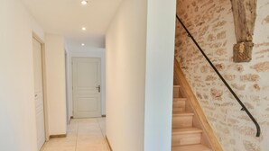 Un pasillo con piso de baldosas, una escalera de madera junto a una pared de piedra, y una puerta al final.