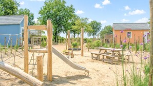 Ein Spielplatz mit Holzkonstruktionen, einer Rutsche, Picknicktischen und einem Gebäude mit einem Ziegeldach.