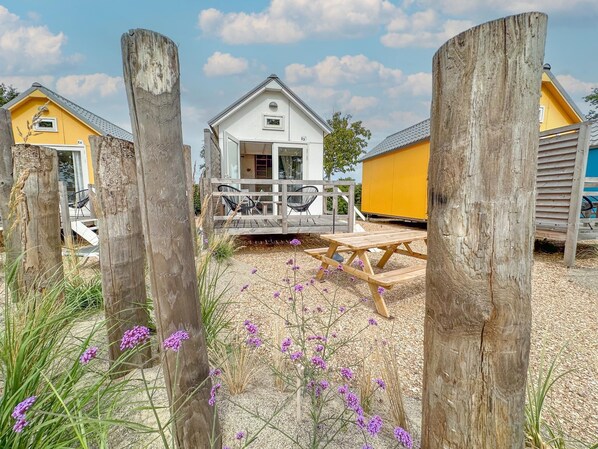A wooden deck with a picnic table, a white house with a porch, and a yellow building in the background. - Detached beach house, quiet location (Ellemeet)