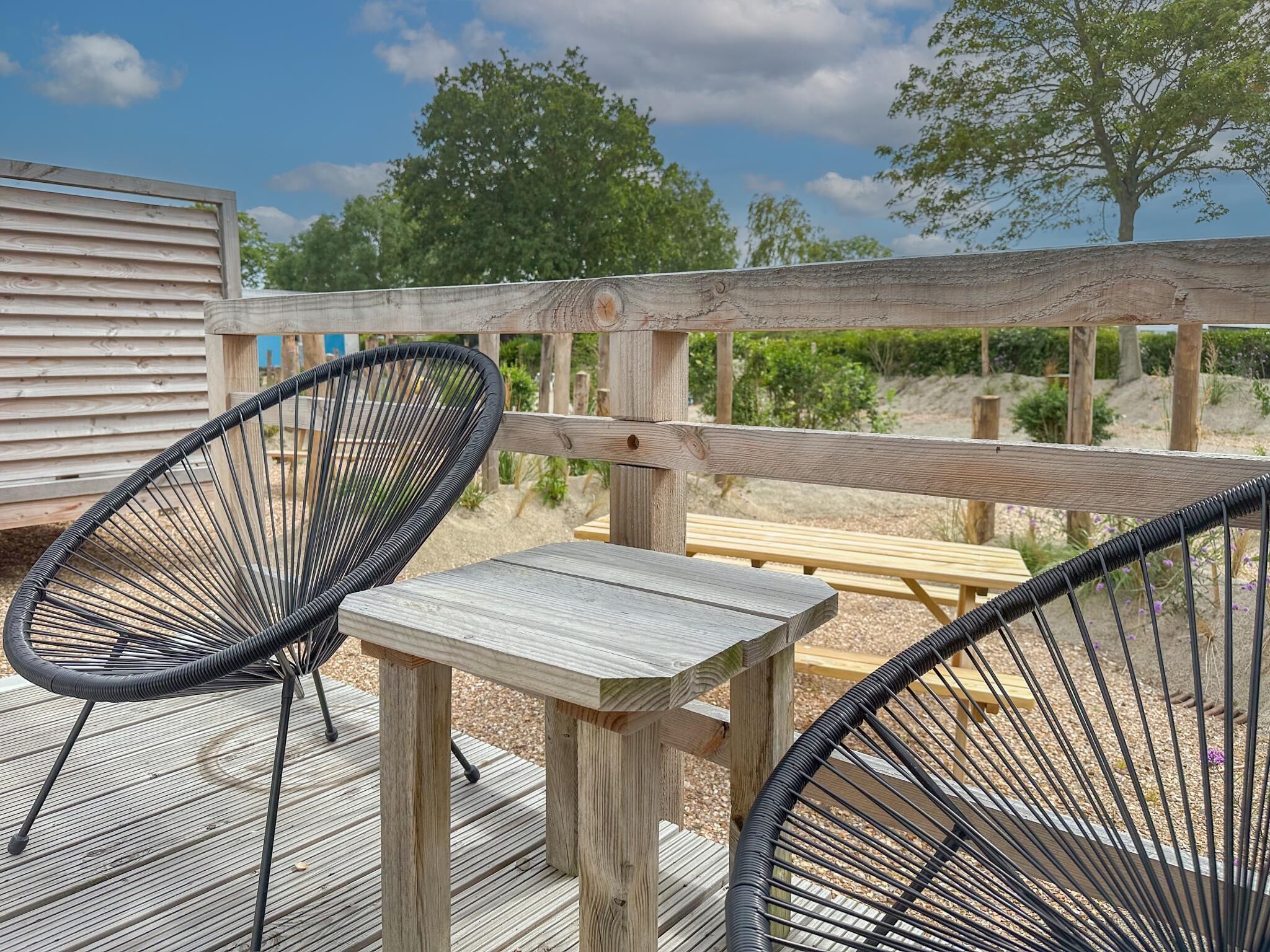 A wooden deck with two black wicker chairs and a small table, surrounded by a wooden fence and trees.