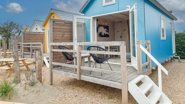 Eine blaue Holzhütte mit einer Holzterrasse, einer Glastür und einer kleinen Sitzgelegenheit im Freien.