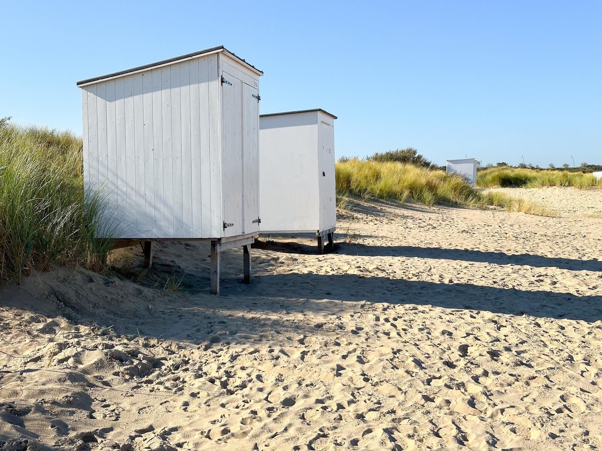 En sandstrand med to hvide bygninger, muligvis strandhytter, uden synlige vinduer eller døre.