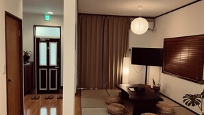 Una stanza con pavimento in legno, un tavolo scuro con una ciotola, cuscini da pavimento intrecciati, una lampada da terra, una TV a schermo piatto e un'ampia finestra con persiane.