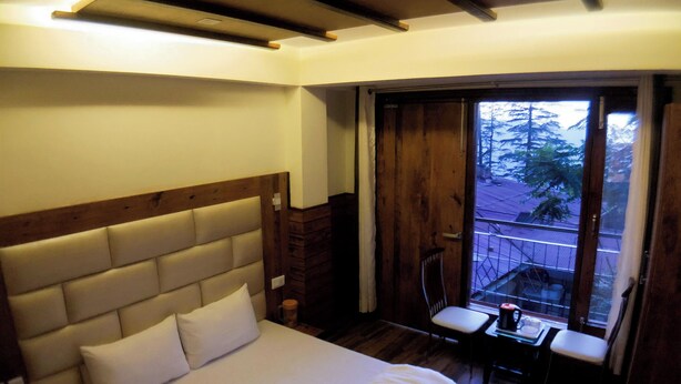 A bedroom with a large bed, wooden headboard, a small table with a chair, and a window with a view of trees. - Backwoods B&B Mall Road Shimla (Shimla)