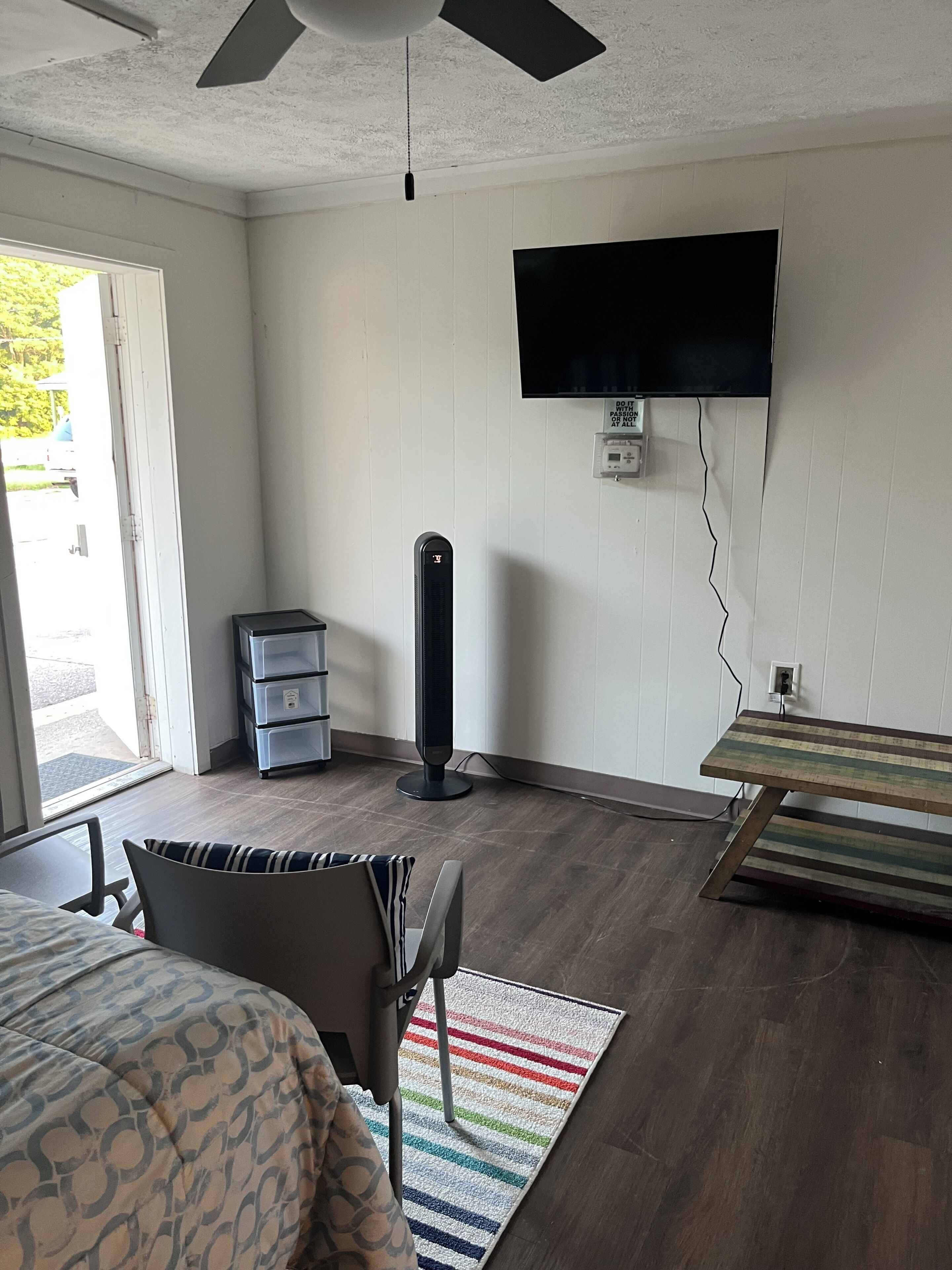 A hotel room with a bed, a desk, a chair, a standing fan, a TV mounted on the wall, and a window with a view of greenery.