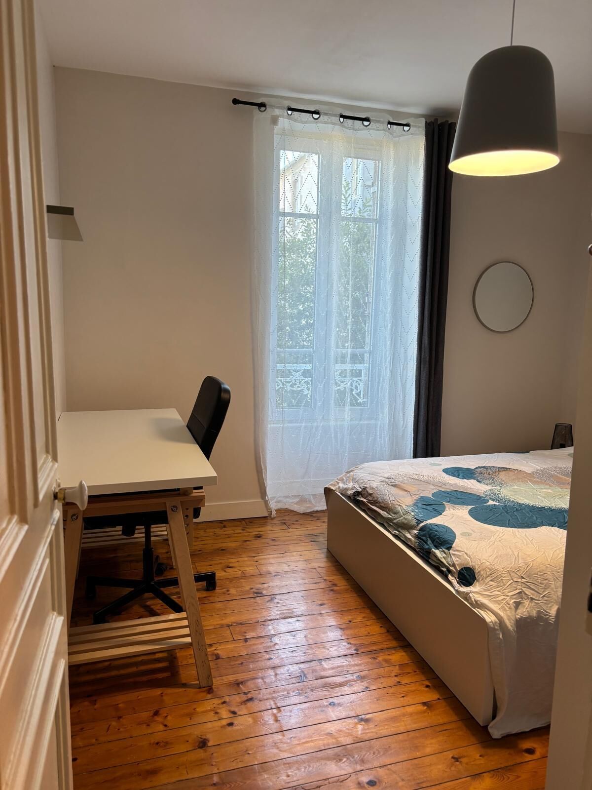 Une pièce comprenant un lit, un bureau, une chaise, un luminaire suspendu et une fenêtre avec des rideaux.