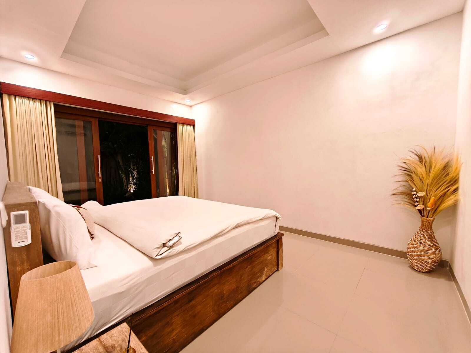A bedroom with a bed, a nightstand, a lamp, a window with curtains, and a decorative vase.